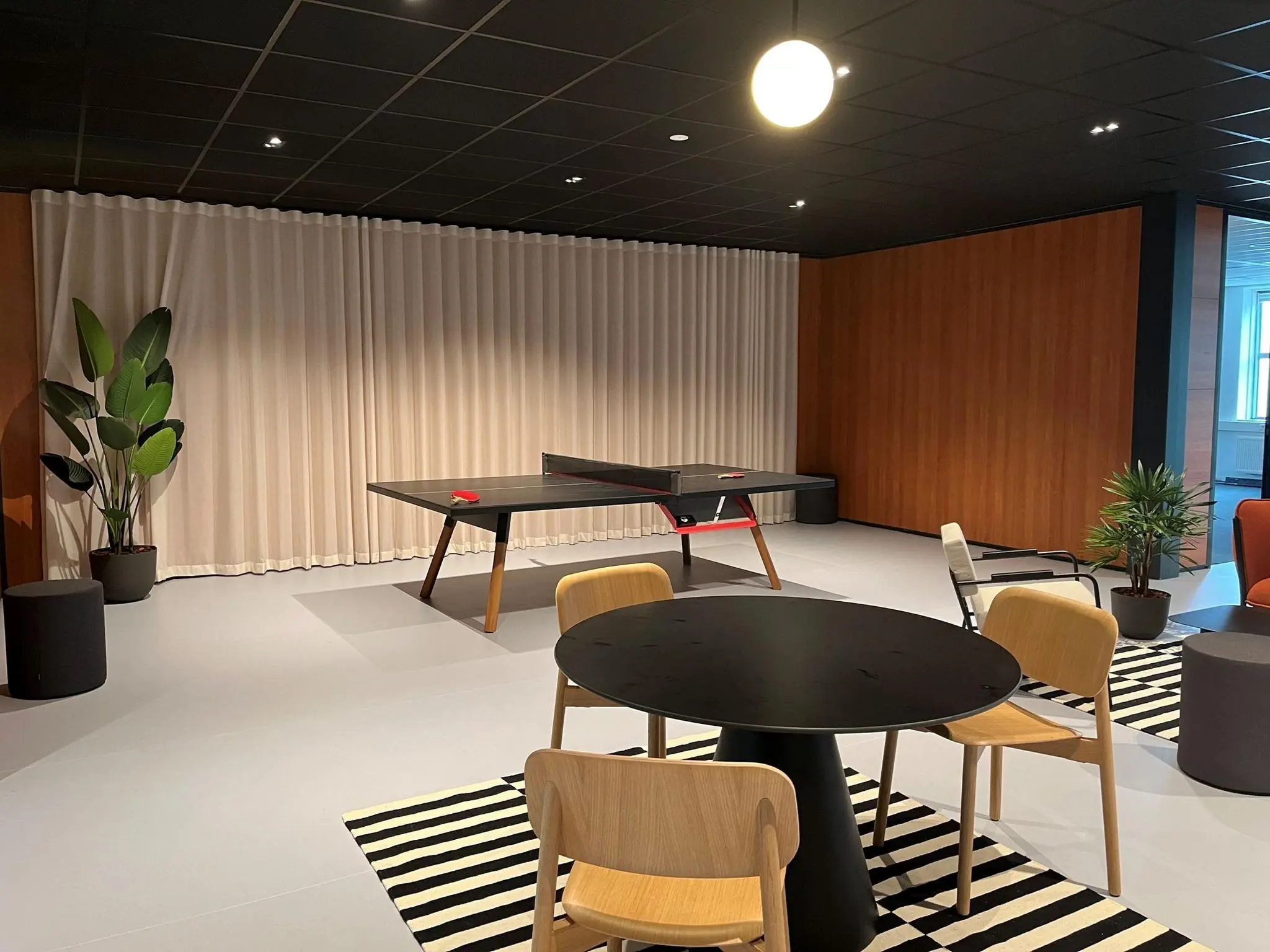 Moderne ontspanningsruimte met een tafeltennistafel, ronde zithoek en planten op een gestreept tapijt.